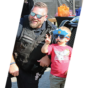 An officer of Tulalip Tribal Police kneeling next to a young toddler boy wearing a stick-on mustache and a cutout police hat. 