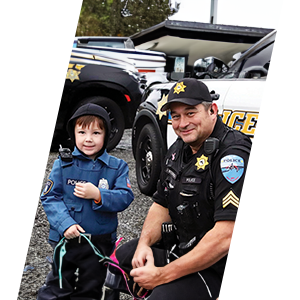 An officer kneeling next to a young boy wearing a child's badge at a community event with patrol cars surrounding them. 